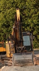 Gąsienicowa koparka z dużą łyżką wysuniętą na pierwszy plan na miejskim placu budowy. W tle widać kabinę operatora, ramię maszyny oraz gęstą ścianę zielonych drzew. © tomo81