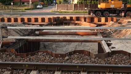 Głęboki wykop budowlany zabezpieczony metalowymi ściankami szczelnymi, zlokalizowany tuż obok torowiska. W tle widoczny jest plac budowy, rury rozporowe oraz fragment dźwigu przy drodze. © tomo81