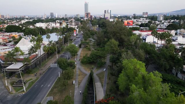 Guadalajara, Mexico - November 16 2025: Backward drone footage captures Lazaro Cardenas Linear Park, an urban street, gas station, Sanborns, and Guadalajara city