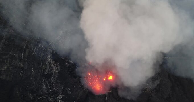 Mount Yasur volcano actively erupting, spewing glowing red lava and thick plumes of white and gray smoke from its crater on Tanna Island, Vanuatu, viewed from an aerial drone view perspective