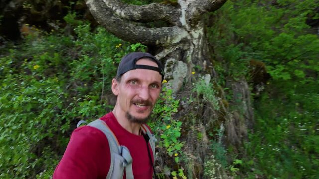 Midground portrait of a bearded man wearing a backward cap and backpack straps, pausing beside a mossy tree trunk in a sun-dappled birch and pine clearing. Lush green undergrowth