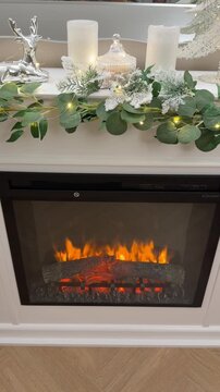Vertical close up of a white wooden fireplace with burning electric insert, decorated for Christmas with a green eucalyptus garland and fairy lights. HD.