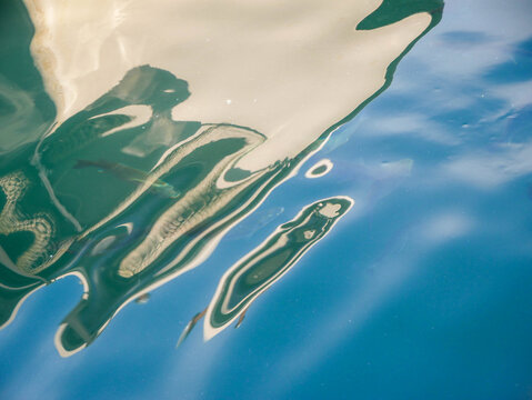 Abstract close-up of a white boat hull reflection distorted by ripples in turquoise-blue marina water, Budva, Montenegro. Fluid painterly shapes in teal, cream and deep blue. Creative abstract