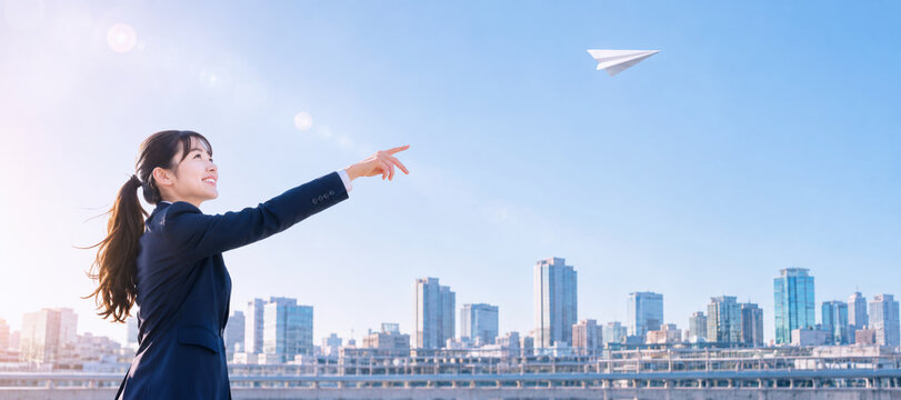 都市の空を背景に紙飛行機を飛ばす女性会社員の明るいポートレート