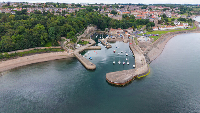 Aerial view of historic Dysart Harbour with boats docked by stone piers, surrounded by lush green park and coastal town in Fife, Scotland.