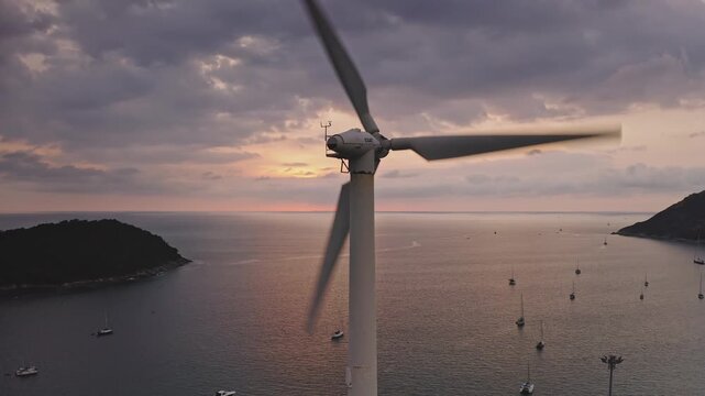 Wind turbine spins at colorful pink sunset over turquoise Phuket waters, sailboats anchored near a lush island, symbolizing clean, renewable energy and coastal eco tourism. Aerial view drone shot