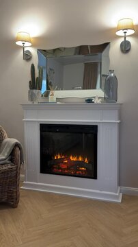 Vertical steady shot of a beautifully crafted white wooden fireplace with a burning electric insert. Elegant living room decor with a wavy mirror and lamps. 4K.