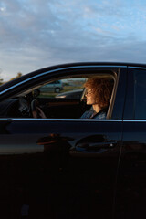 Naklejka premium A woman sits in the driver's seat of a dark car, profile visible through the window as evening light glints on the panel. A quiet moment of travel and personal reflection.