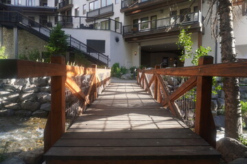 Fototapeta premium A eye-level, straight-on shot looking across a rustic wooden footbridge with cross-braced railings. The bridge spans over a small rocky stream or creek, leading toward a modern alpine resort complex o