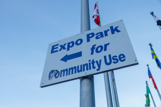 A blue and white Expo Park for Community Use directional sign points the way beneath international flags and a clear sky near False Creek.