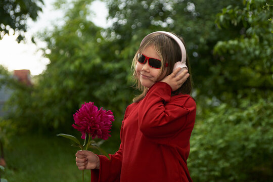 Portrait of a happy girl in headphones with a burgundy peony in her hand against a picturesque green garden background.