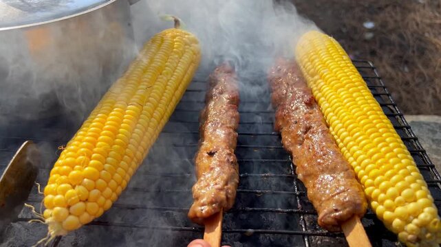 Sweet corn cobs and traditional Turkish Adana kebab skewers cooking on hot outdoor charcoal grill with thick rising smoke.