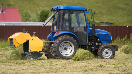 Obraz premium A mini hay baler works in cramped conditions, baling small bales. The outskirts of a village are in the background.