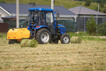 Obraz premium A farmer harvests hay on a small plot on the outskirts of a village. He works with a mini tractor and a towed mini baler.