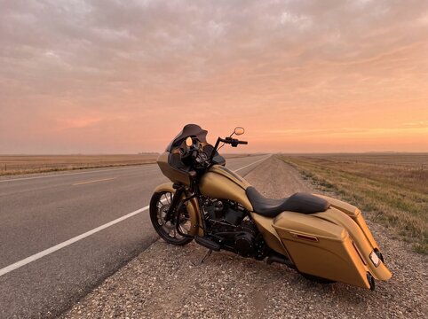 A long, satin gold bagger motorcycle parked at the edge of a flat, straight midwestern highway that vanishes into the horizon, professional photography, no logo.