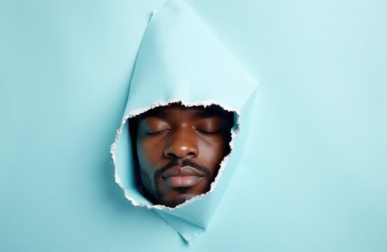 Young black man peeks through torn paper with eyes closed. Peaceful face emerges from hole in light blue background. Calm expression, serene mood, introspective moment.