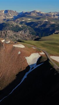 Vertical Screen: Beartooth Highway in Montana USA curves through alpine terrain with snow patches on rugged slopes and distant mountain peaks under clear blue summer sky.