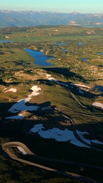 Vertical Screen: Beartooth Highway in Montana USA with winding mountain road cutting through alpine terrain with snow patches and blue lakes dotting the green valleys below.