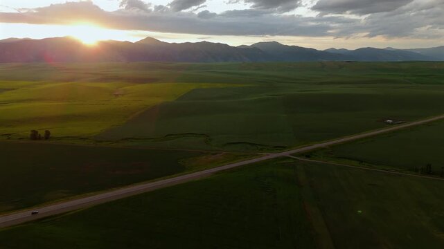 Aerial Montana USA view captures stunning golden hour sunset illuminating expansive green farmland and rolling hills. Dramatic clouds drift over mountain silhouettes as rural roads cross valley floor.