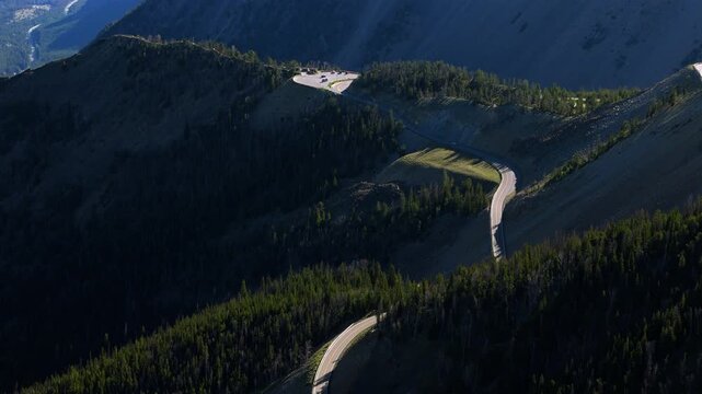 Aerial of Beartooth Highway in Montana, USA showing winding road cutting through forested mountain ridges. Scenic drone view captures serpentine curves and parking area with dramatic alpine terrain.