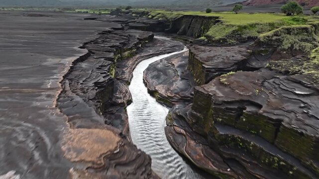 Meandering river flows through an ancient volcanic canyon on Tanna Island, Vanuatu, carving intricate patterns into the hardened lava flows and black sand landscape. Aerial view fast drone flight