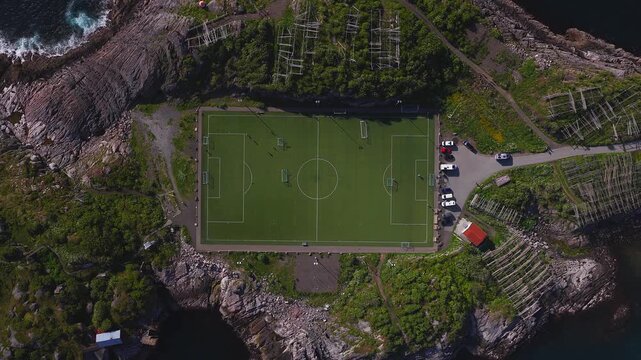 Aerial top down view of Henningsvaer pitch in Lofoten, Norway. Players move, cars arrive, waves break. Turf field, hjell, and red boathouses sit on granite under summer light.