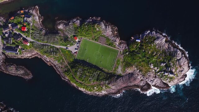 Aerial top down view shows Henningsvaer football field on a rocky promontory in Lofoten, Norway, with coastal houses, hjell racks, parked cars, surf, and sea.