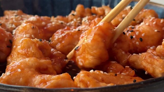 A person uses chopsticks to pick up and enjoy a chicken wing coated in sweet and sour sauce with sesame seeds