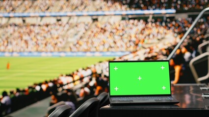Close-up of isolated green screen on laptop in football stadium stands during a live sport event, soccer crowd celebrating in the background with scarves, stadium seats and bright arena lights. © DC Studio