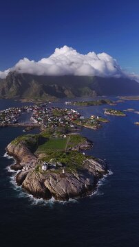 Aerial view of Henningsvaer football pitch in Lofoten, Norway, with lighthouse, skerries, docks, boats, village streets, and jagged mountains under rolling clouds.