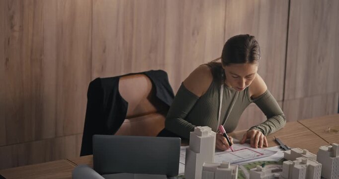 An architect works on building plans while sitting at a desk filled with models in an office environment