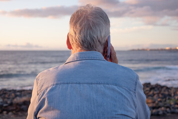 Fototapeta premium Senior man with gray hair standing on the beach at sunset, holding a smartphone to his ear and looking toward the ocean, relaxed and connected during retirement