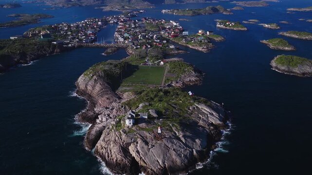 Aerial view of Henningsv&aelig;r Football Soccer Stadium. Bright green turf pitch on a rocky islet, stockfish racks, skerries, boats, cars, and cloud capped peaks in summer light.