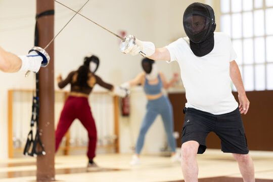 Elderly man in a protective helmet holds a rapier and participates in a training match in a fencing club