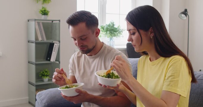 eating takeout salad together at home. On a sofa, man and woman smile while enjoying fresh bowls in living room having lunch. Healthy takeaway meal at home. Delivery service concept