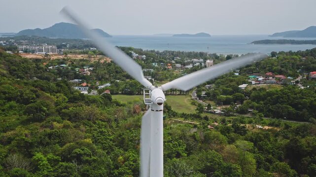 Wind turbine spinning its blades, generating clean, renewable energy overlooking a tropical coastline, lush green landscape, and residential areas of Phuket, Thailand. Aerial view drone shot