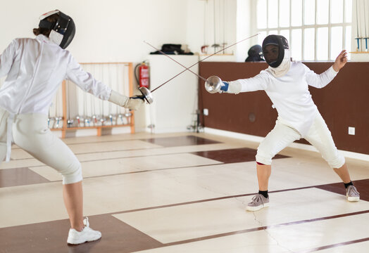 Athletes in protective equipment and helmets conduct a fencing training match in the gym. People at a group fencing lesson practice with rapier