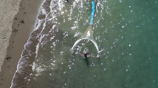 Aerial drone view of a fisherman wading in shallow coastal waters while operating a traditional fishing net along a sandy beach.