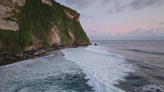 Coastal mountain cliffs covered in lush green vegetation meet ocean waves crashing on the shore during colorful sunset, showcasing a tropical landscape and natural beauty of Bali island, Indonesia