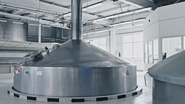 Large stainless steel tanks for beverage fermentation and brewing standing inside a clean, modern production hall of an industrial brewery factory with nobody around in a quiet working environment