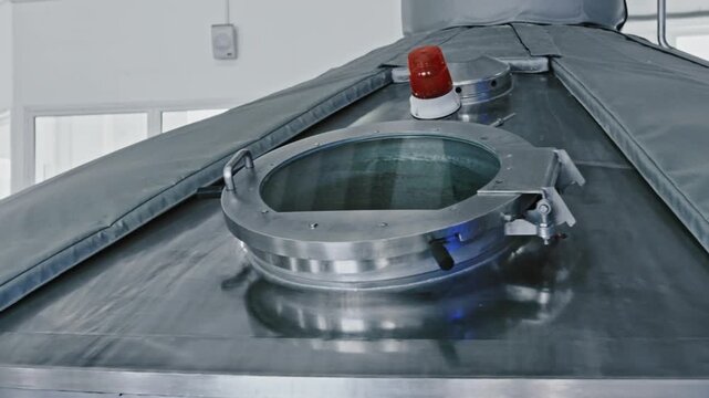 Automated manufacturing process inside a modern brewery with a large, shiny stainless steel beer fermentation tank featuring an open hatch and a flashing red warning light on top
