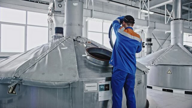 Professional industrial employee in a blue uniform opening the round hatch of a large stainless steel brewing vat inside a modern beverage production factory for quality control