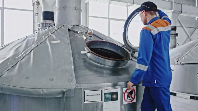 Male worker in a blue uniform opening a large stainless steel vat, inspecting the industrial equipment during the brewing process inside a modern beverage manufacturing plant