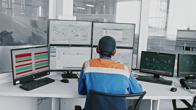 Male engineer working in a modern control room, sitting at a desk and supervising production processes on several computer monitors displaying diagrams, charts, and data for process control