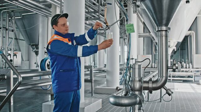 Industrial brewery worker in a uniform taking a beer sample from a large stainless steel tank for quality control, checking the beverage's color and clarity during the production process