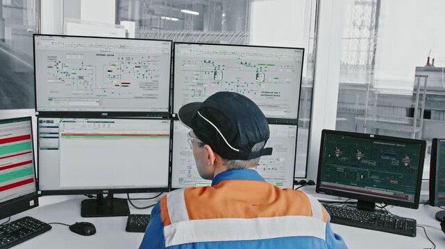 Industrial engineer in a control room, sitting at a desk with multiple monitors, supervising automated manufacturing processes and checking technical data on computer screens for quality control