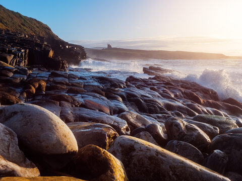 Sunlight glints off wet, rounded stones along the shoreline. Waves crash against the rocks with force and rhythm. Castle Classiebawn in the background. County Sligo, Ireland. Travel and tourism.