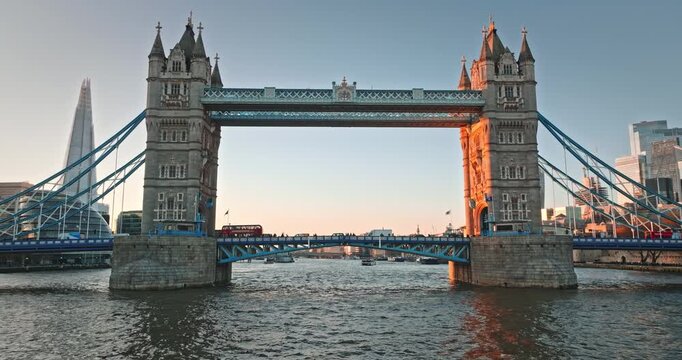 Tower Bridge, a historic suspension bridge, crosses the River Thames with boats and a red bus during sunset, featuring The Shard and London cityscape skyline. United Kingdom travel destination