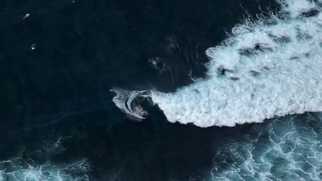 Surfer riding powerful deep blue ocean wave, creating white foam as other surfers wait in the water, captured from an aerial perspective over the Bukit Peninsula in Bali. Active sports and recreation