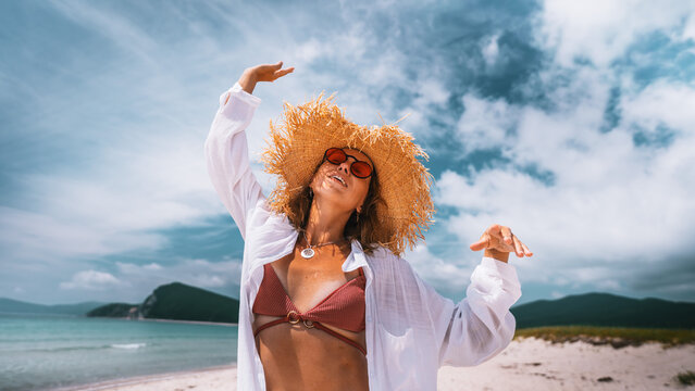 Elegant woman in trendy swimwear and oversized straw hat poses on beautiful beach. Luxury vacation atmosphere, fashionable accessories and coastal scenery inspire modern travel content. Fashion